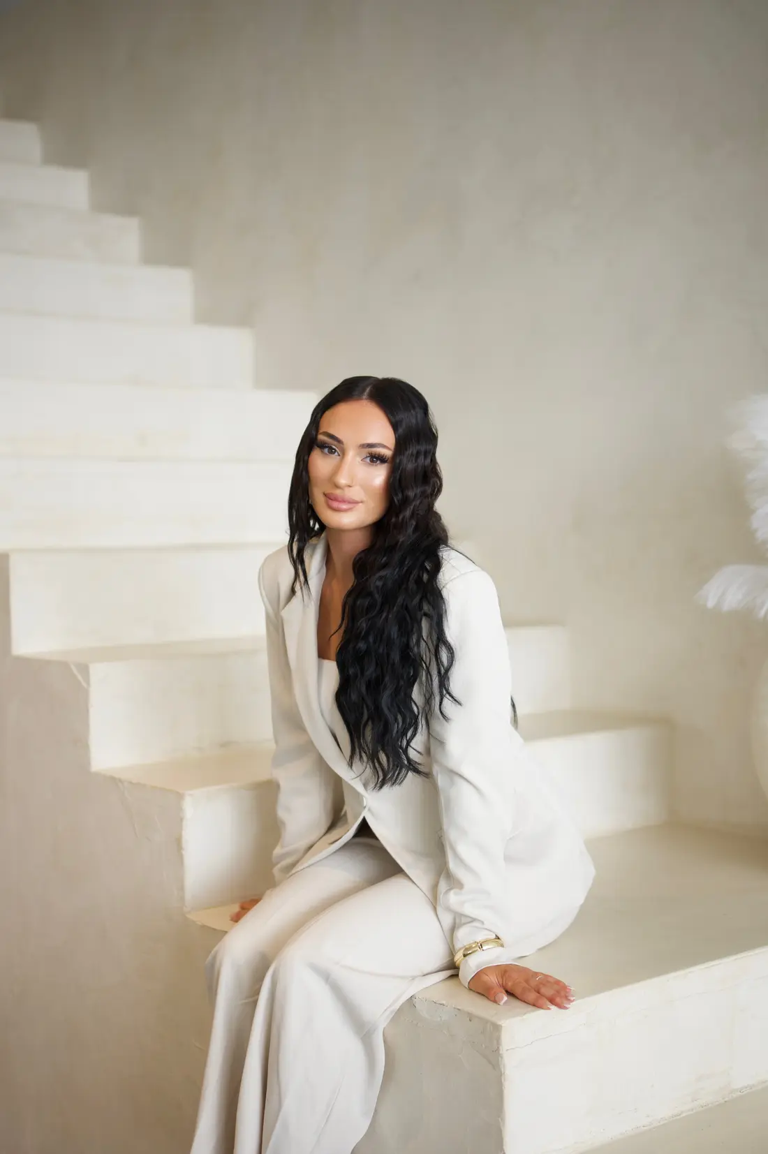 Leyla, Inhaberin von LEYLABEAUTY in Troisdorf, sitzt entspannt auf einer hellen Treppe im cremefarbenen Anzug
